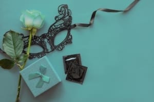 Love, passion, sex romance flat lay, mock up with red rose, black lace carnival mask, gift box and two condoms on red background. Safe sex, dating concept. Top view